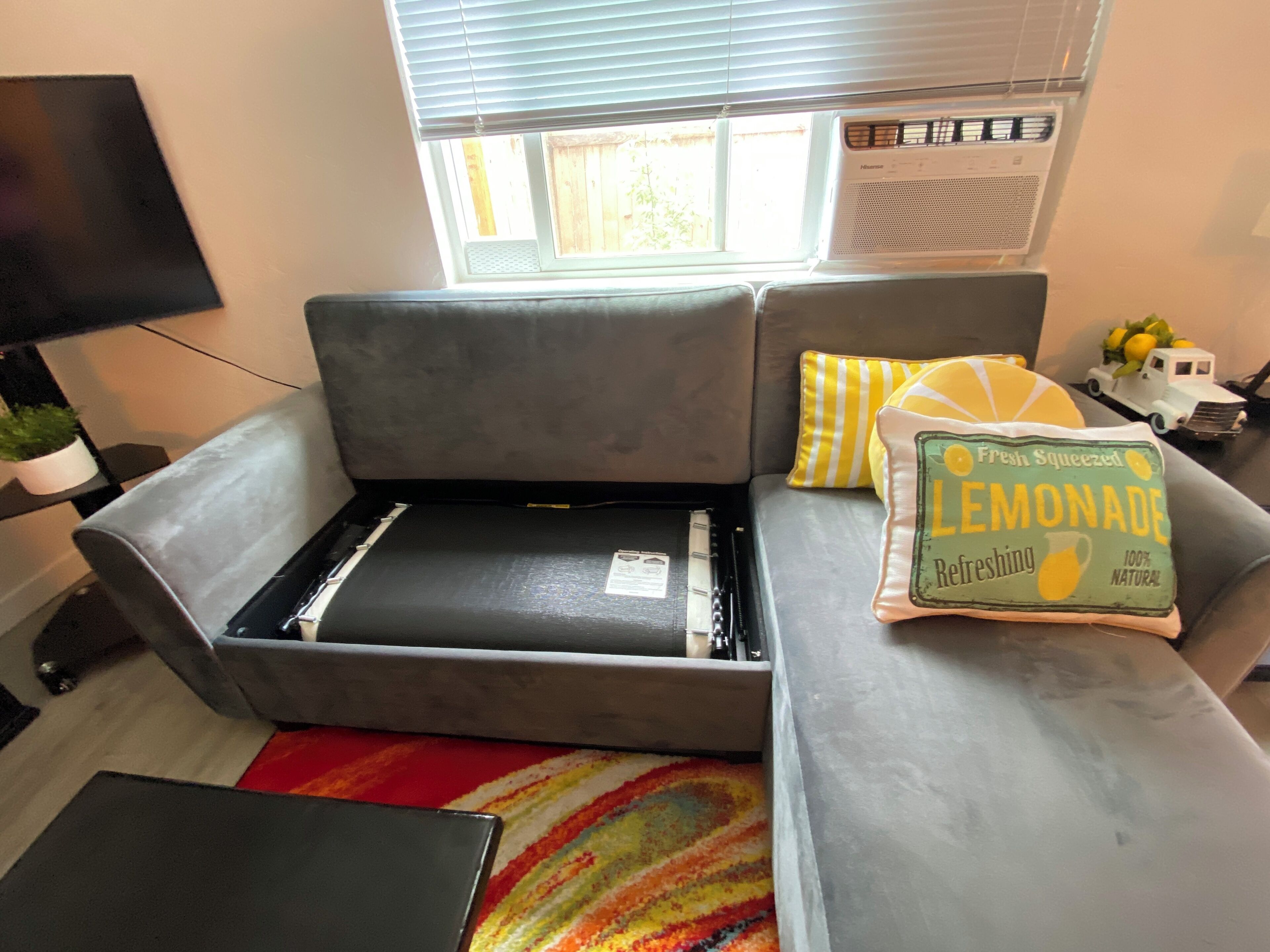 A grey sofa with a built-in table, a small table with a lamp, a colorful rug, and a pillow with the text 'Fresh Squeezed Lemonade Refreshing 100% Natural.'