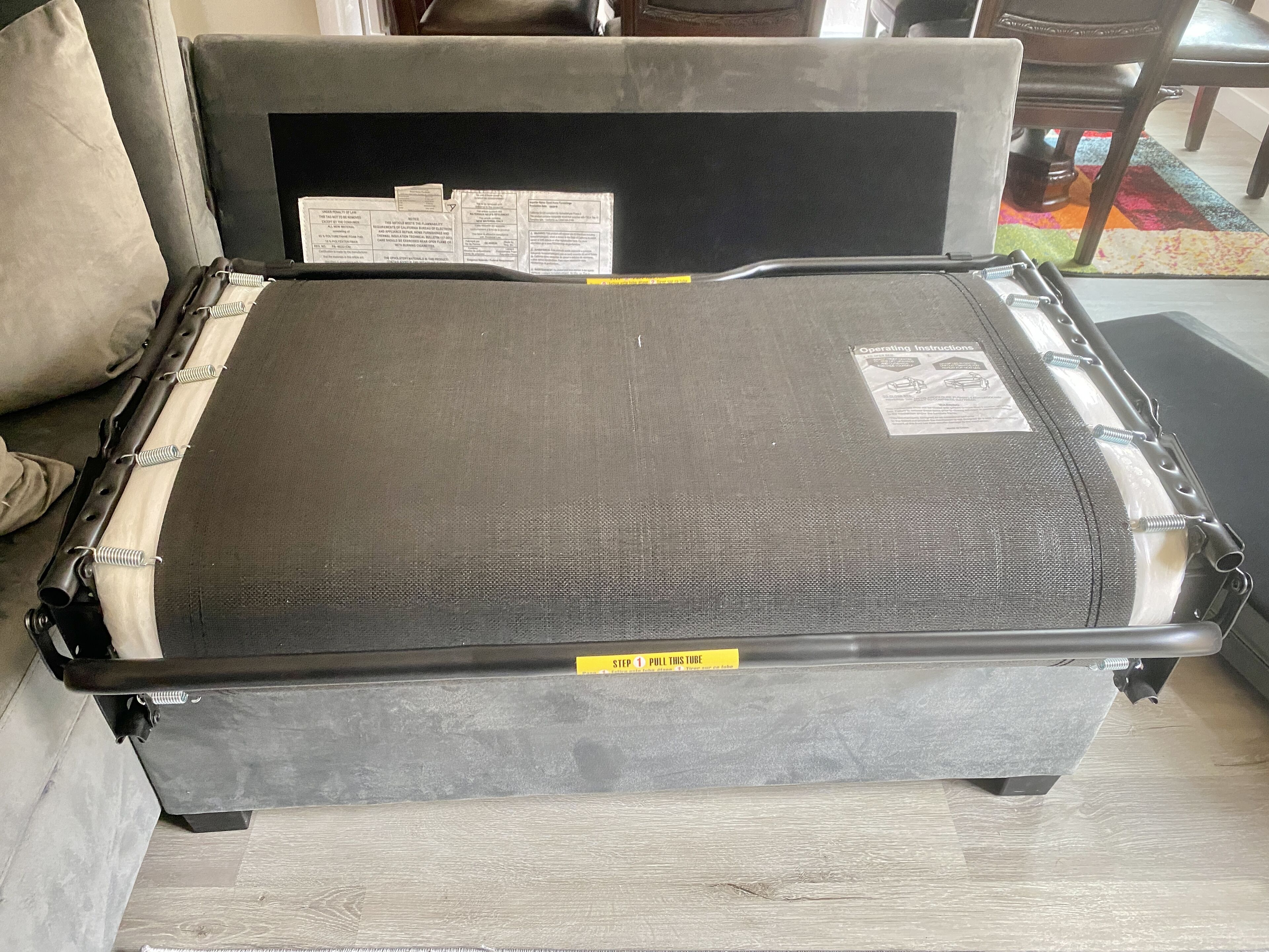 A bed with a grey headboard and footboard, covered with a grey mattress and a protective plastic sheet. There is a yellow tag on the footboard with the text "STEP 1 PULL THIS TUBE".