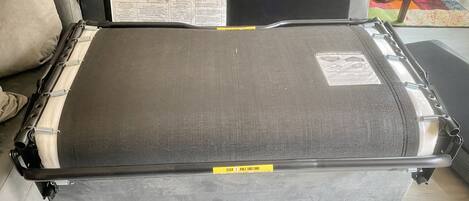 A bed with a grey headboard and footboard, covered with a grey mattress and a protective plastic sheet. There is a yellow tag on the footboard with the text "STEP 1 PULL THIS TUBE".