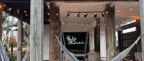 A wooden structure with a hammock, wooden chairs, and a neon sign reading "Gulf Shares".