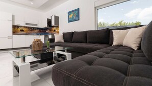 Un salon moderne avec un canapé d’angle gris foncé, une table basse en verre et une cuisine en arrière-plan.
