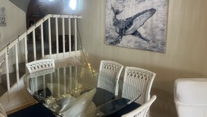 Un comedor con una mesa de vidrio, sillas de mimbre blanco, una escalera con barandilla y un cuadro de una ballena colgado en la pared.