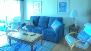 Una sala de estar con un sofá azul, una mesa de centro, una lámpara y un sillón con un cojín de temática playera.