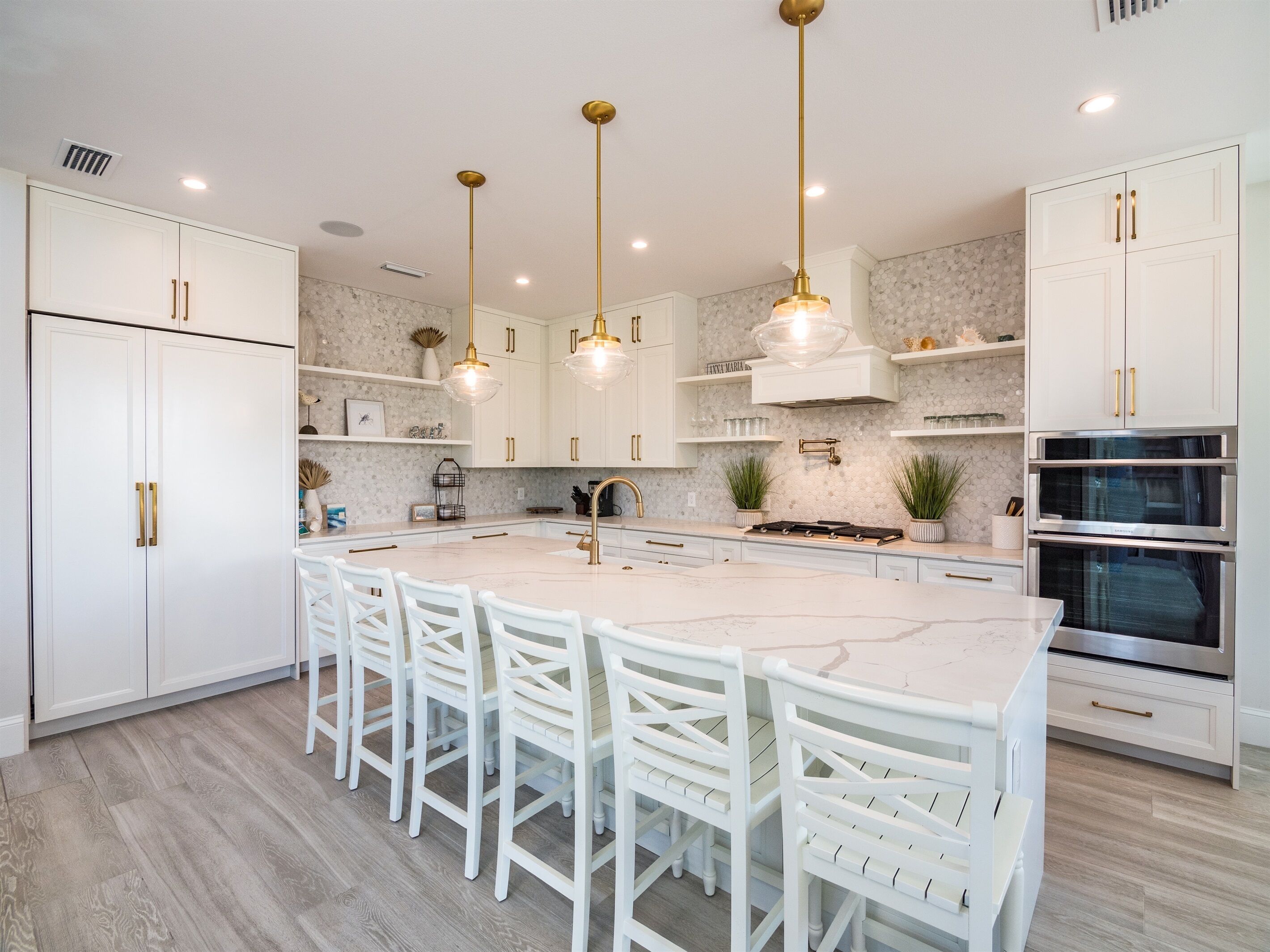 A modern kitchen with white cabinetry, a large island with bar stools, pendant lighting, and built-in ovens.