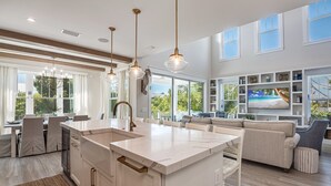 A modern kitchen with a central island, pendant lighting, and a dining area with a view of the outdoors. - Paradise Blu by Beach Boutique Rentals (Anna Maria)