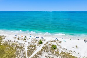 Een strand met helder turquoise water, wit zand en verschillende parasols en strandstoelen.