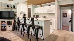 A compact kitchen with white cabinets, a microwave, and a coffee maker. There is a small island with stools and a television on a shelf.