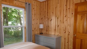 Ein Schlafzimmer mit Holzvertäfelung, einem Bett, einer Kommode, einem Fenster mit Blick auf Bäume und einer Tür.