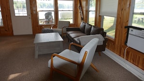 Ein gemütliches Zimmer mit Holzvertäfelung, einer weißen Tür, einem grauen Sofa, einem Holztisch und Blick auf einen See durch große Fenster.