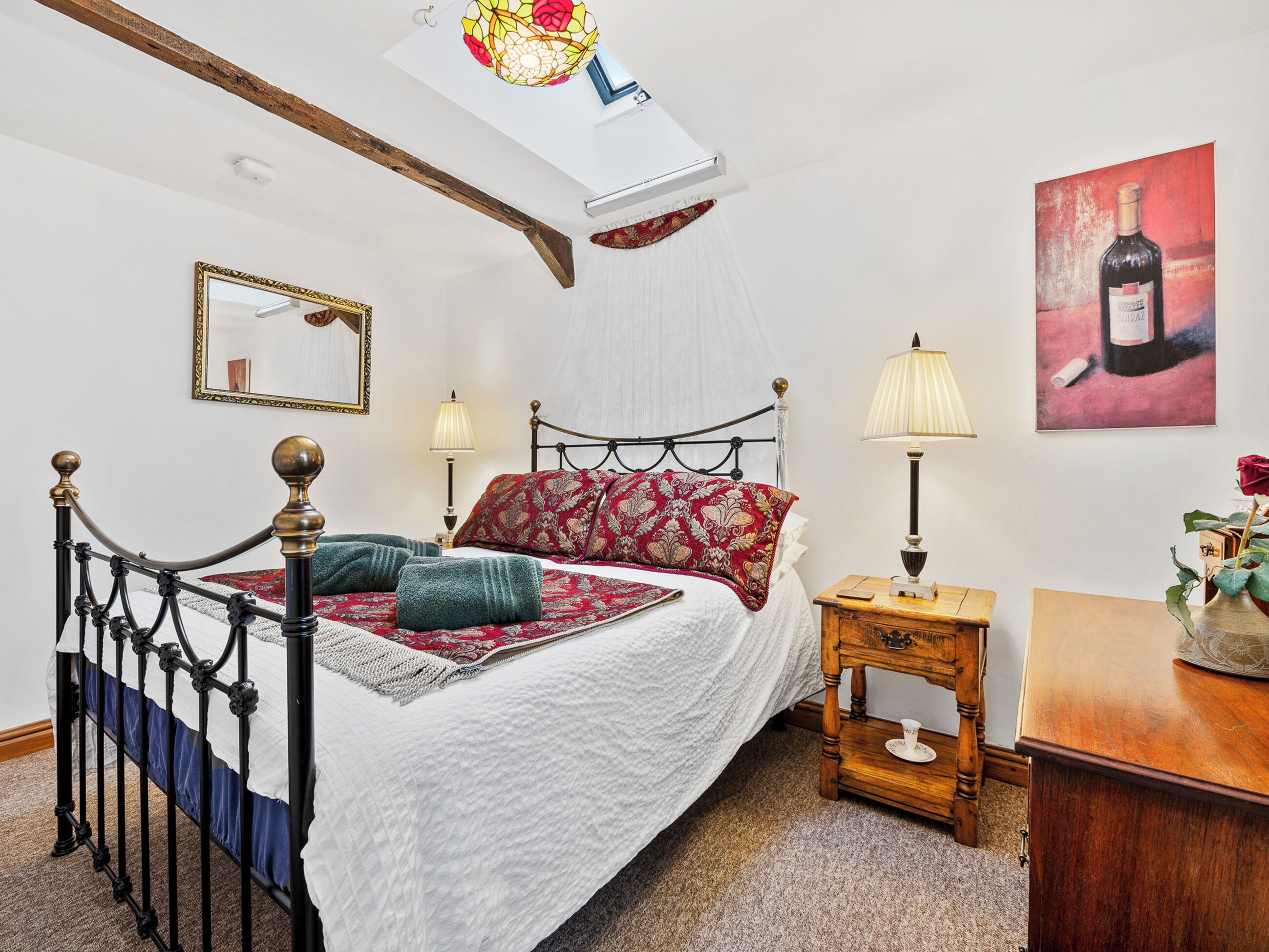 A bedroom with a bed, bedside lamps, a mirror, a wooden nightstand, and a painting.