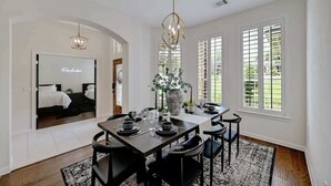 Un espace repas avec une grande table dressée pour six personnes, un lustre et une vue sur une chambre à coucher, visible à travers une arche.