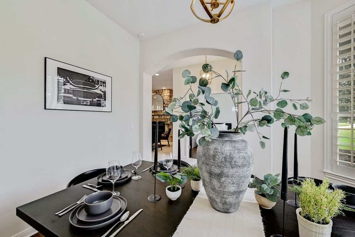 Un coin repas avec une table en bois foncé, des assiettes noires et des couverts en argent. Un grand vase gris contenant des feuilles vertes est placé au centre.