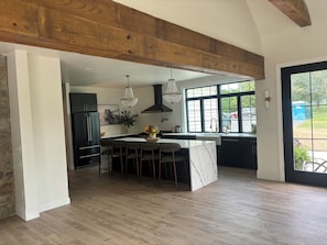 A modern kitchen with a large island, dark cabinetry, and a view through large windows.