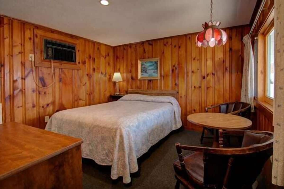 A wooden-paneled room with a bed, a small table, and a chair.