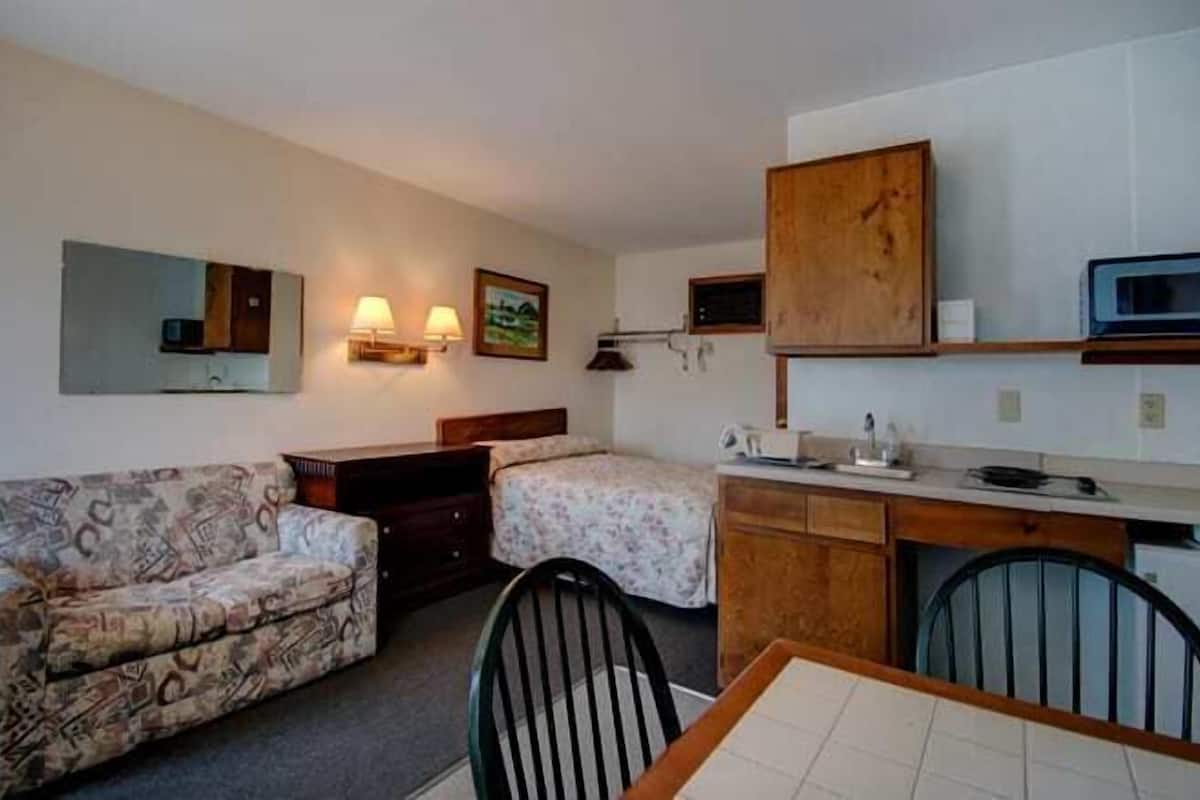 A compact hotel room with two single beds, a sofa, a small dining table, and a kitchenette.