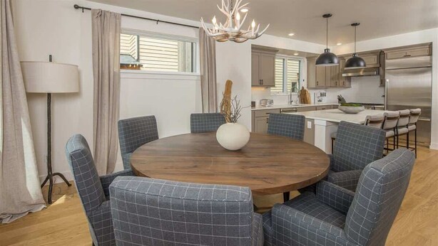 Un coin repas avec une table ronde en bois, entourée de six chaises rembourrées à motifs à carreaux. Un grand vase contenant des branches est posé sur la table.