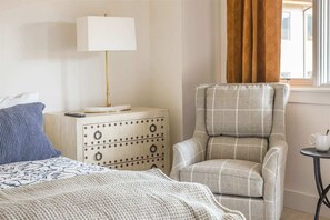Hotellihuone, jossa on sänky, yöpöytä lampun kanssa, tuoli ja pieni pöytä, jossa on kuppi.