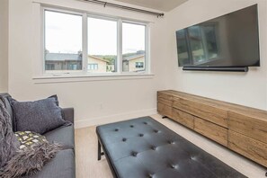 Un salon moderne avec un canapé gris, un pouf noir, un meuble en bois et un grand téléviseur à écran plat fixé au mur.