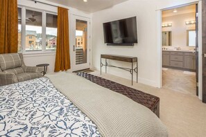 A bedroom with a large bed, a TV mounted on the wall, a bench, and a view of the outside through a window.