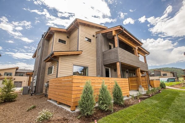 A modern multi-story house with a wooden exterior, a balcony, and a well-maintained garden with small trees.
