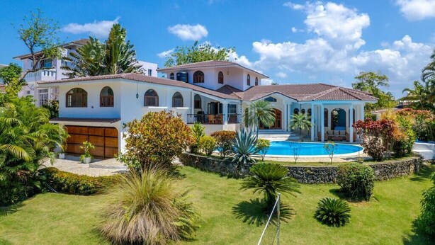 A large, multi-story house with a swimming pool, surrounded by lush greenery and palm trees.