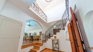 A spacious interior with a staircase leading to a balcony, a dining area in the background, and a ceiling fan.