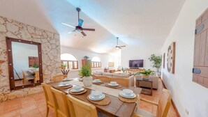A spacious living area with a dining table set for four, a sofa, a TV, and a ceiling fan.