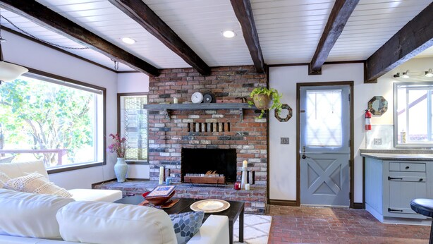 A living room with a brick fireplace, a white sofa, and a kitchen area with white cabinets. - Riverside Home With a Rustic Charm (Washougal)