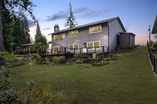 A modern two-story house with a deck, surrounded by a well-maintained garden and tall trees.