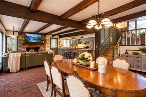 A spacious dining area with a large wooden table, surrounded by upholstered chairs. A fireplace with a TV mounted above it is visible in the background.