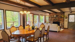 A dining area with a wooden table and chairs, a chandelier, and large windows with a view of greenery. - Riverside Home With a Rustic Charm (Washougal)
