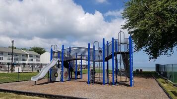 A playground with a slide and climbing structures, surrounded by a fenced area and a building in the background.
