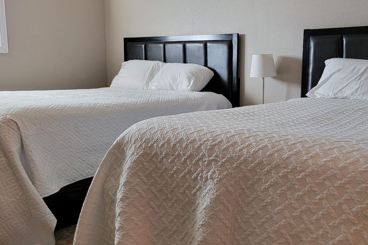 Dos camas con cabeceras de madera oscura, ropa de cama blanca y una lámpara de noche.