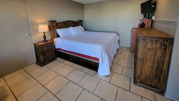 A bedroom with a wooden bed, a nightstand with a lamp, a wooden cabinet, and a television on a stand. - Five Star Inn (Eunice)