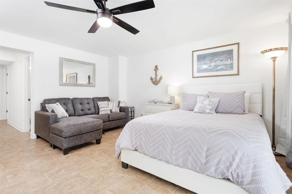 Une chambre à coucher avec un lit, un canapé gris, une table de chevet, un ventilateur de plafond, une lampe fixée au mur et un tableau encadré.