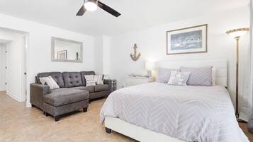 Une chambre à coucher avec un lit, un canapé gris, une table de chevet, un ventilateur de plafond, une lampe fixée au mur et un tableau encadré.