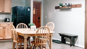 Un comedor con una mesa y sillas de madera, un refrigerador negro, armarios de madera y un pequeño banco.