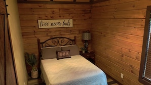 A cozy wooden cabin bedroom with a bed, a nightstand, a lamp, and a sign that says "love never fails". - Bennett Lake Tiny Cabin  (Whitwell)
