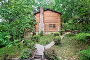Une cabane en bois, entourée d’une végétation luxuriante, avec une cheminée en pierre et un escalier en bois menant à l’entrée.