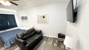 Ett modernt rum med en lädersoffa, en säng med ett blått överkast, ett litet bord med en lampa och en väggmonterad TV.