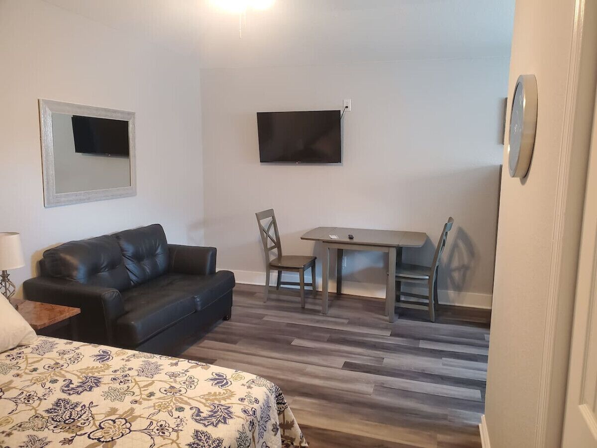 A room with a bed, a black leather sofa, a dining table with chairs, a flat-screen TV mounted on the wall, and a patterned bedspread.