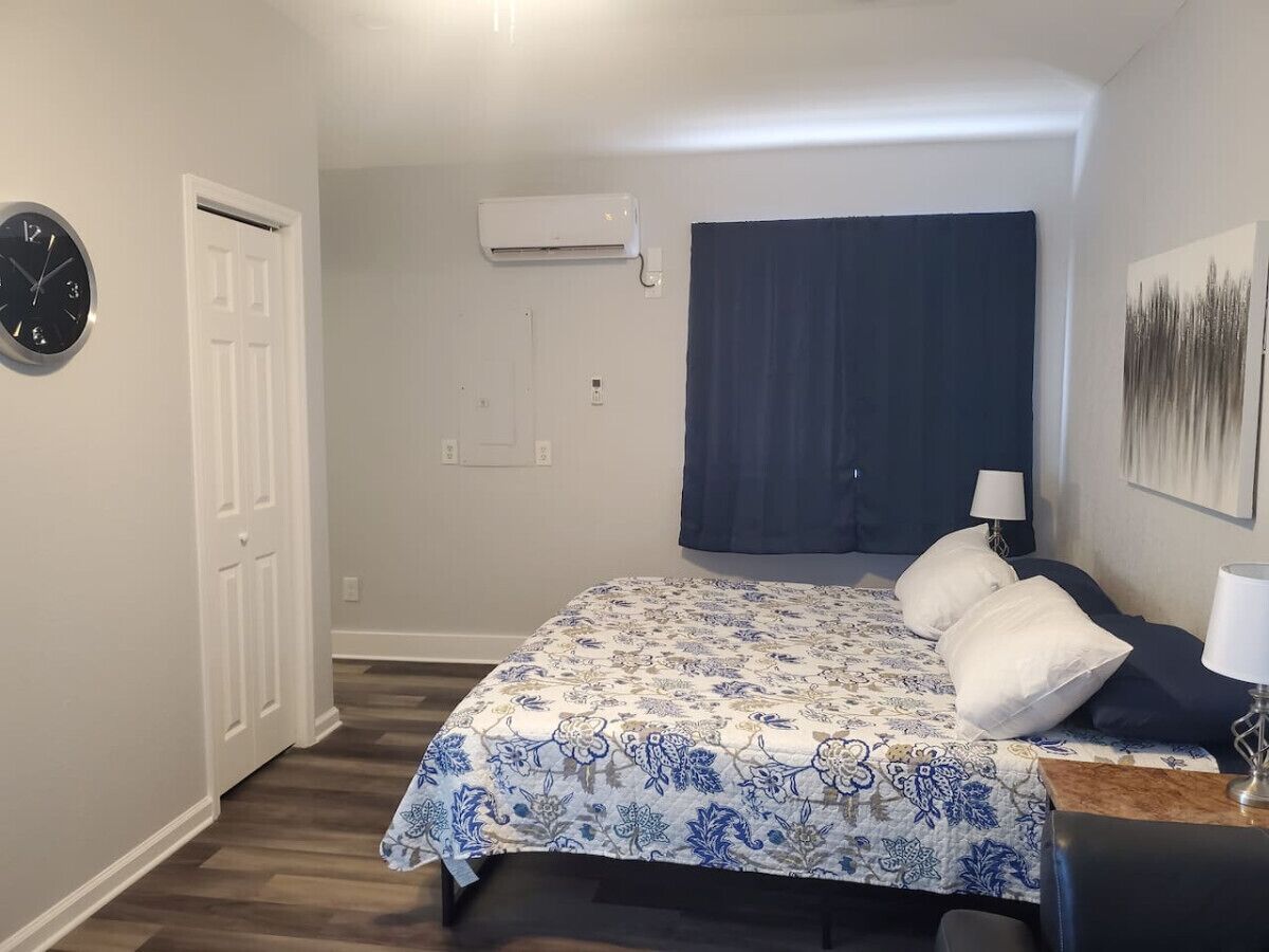 A bedroom with a bed, a wall clock, an air conditioner, a window with a dark curtain, and a painting on the wall.