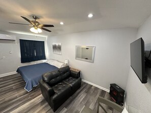 Une pièce comprenant un lit, un canapé en cuir noir, un ventilateur de plafond, une télévision et une fenêtre avec des rideaux.