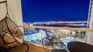 A balcony with a hanging chair and a table, overlooking a cityscape at night with illuminated buildings and a pool area. - MGM Signature 1BR 2BA Wet Republic, Top Golf & Strip Views / NO Resort Fee (Las Vegas)