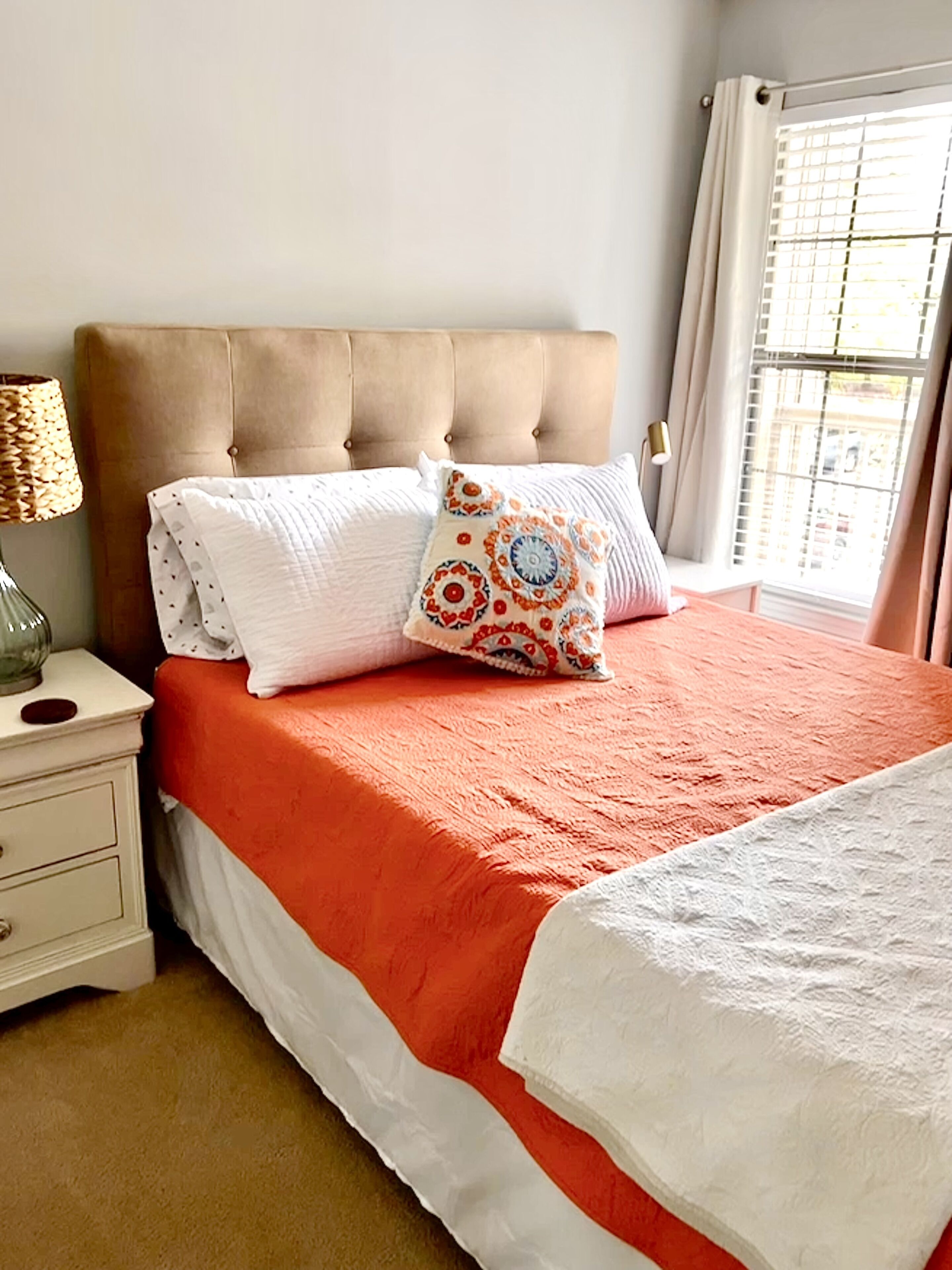 Een slaapkamer met een gestoffeerd hoofdeinde, een bed met een oranje sprei, witte kussens en een bedlampje.