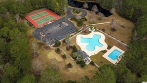 An aerial view of a property with a tennis court, a swimming pool, and a parking area surrounded by trees. - Beautiful 2 bed 2 bath Corner Unit with golf course view-5 minutes from beach (Pawleys Island)