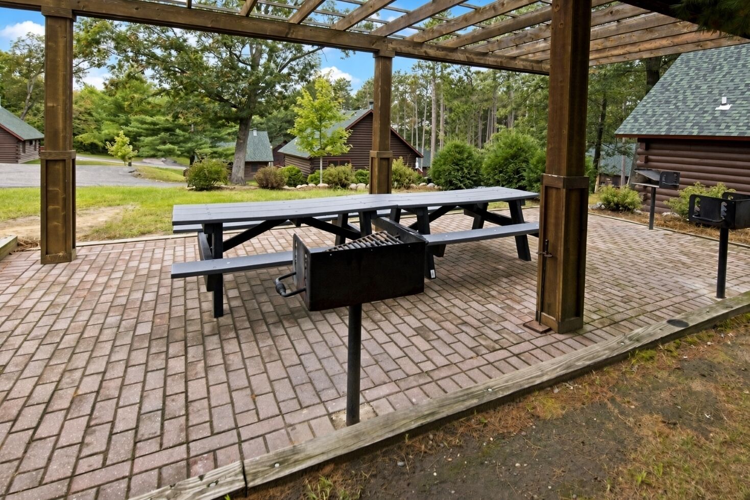 A covered picnic area with a table and grill, surrounded by a wooden pergola, with a view of cabins in the background.