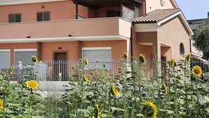 A two-story building with a balcony, a gated entrance, and a row of sunflowers in the foreground. - C006 - Camerano, new three-room apartment with garden (Camerano)