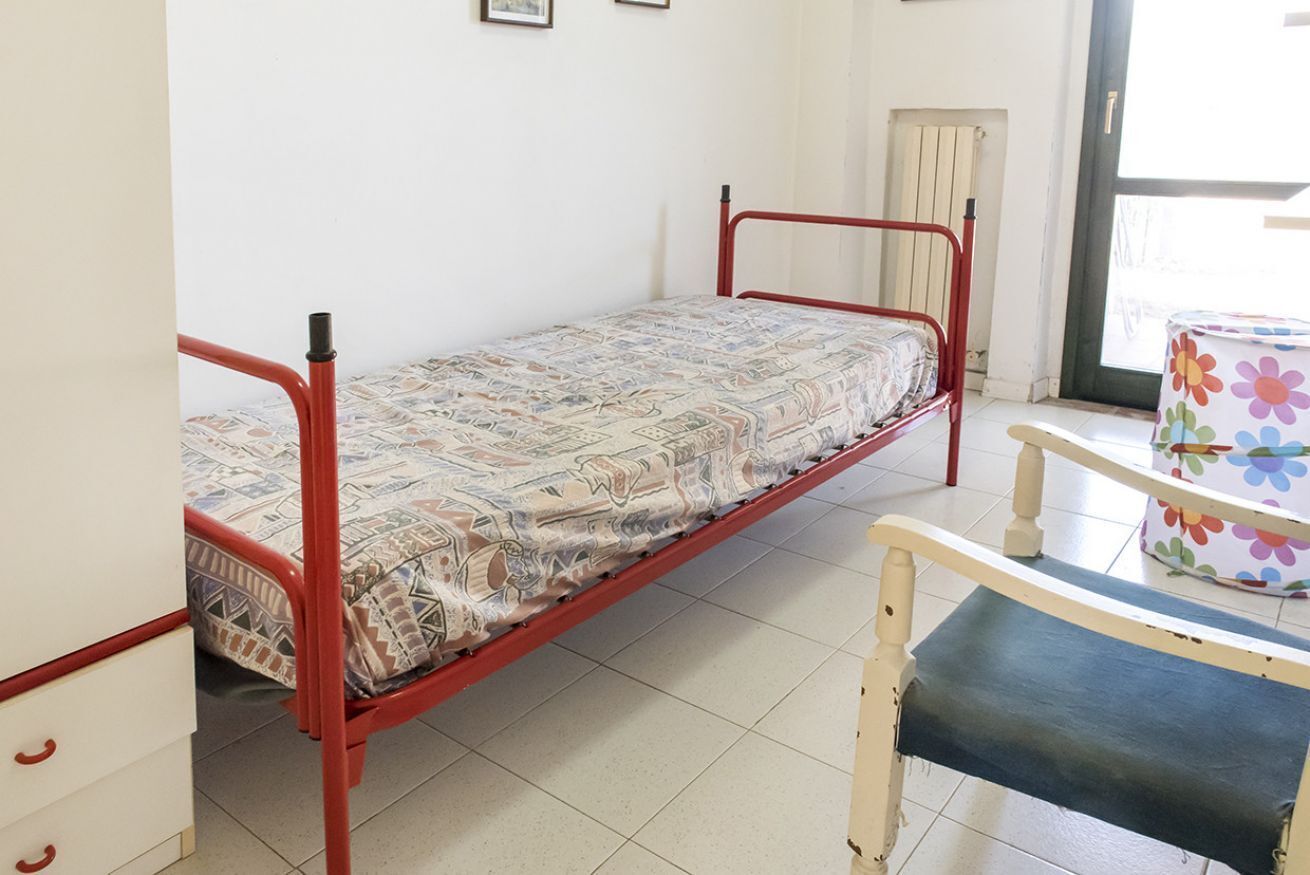 Un lit simple avec un matelas à motifs, une chaise en bois avec un coussin à fleurs et une fenêtre donnant sur l’extérieur.