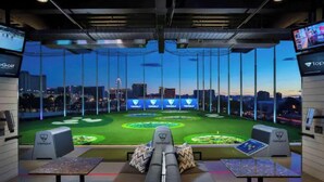 Sala de simulador de golf con múltiples pantallas mostrando un campo de golf, una vista del horizonte urbano al anochecer y un panel de control para el simulador.